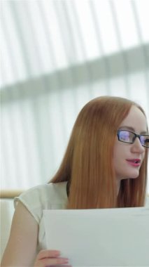 Kendine güvenen kızıl saçlı ve şık gözlükleri olan bir kadın, şık giyimli bir adamla önemli belgeleri tutkuyla tartışıyor ve takım çalışmasının dinamik atmosferini teşvik ediyor. Arka planda, iki odaklanmış profesyonel, bir erkek ve bir kadın,