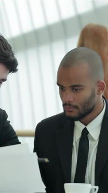 Biri siyah saçlı, diğeri tıraşlı iki kendine güvenen işadamı, modern bir ofis masasında profesyonellik ve hırs dolu belgeler üzerinde yoğun bir şekilde tartışıyorlar. Ciddi ifadeleri işbirliğine olan bağlılıklarını yansıtırken,