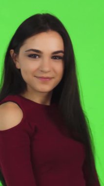 Uzun, dalgalı esmer saçlı çekici bir kadın kamerayı neşeyle işaret ediyor. Parlak gülüşü canlı yeşil arkaplanı aydınlatıyor. Bulaşıcı enerjisi ve kendine güveni davetkar bir atmosfer yaratarak izleyicileri iletişim kurmaya teşvik ediyor.