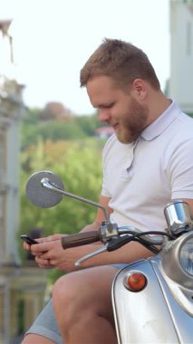 İyi bakımlı sakallı neşeli bir genç adam şık scooterına binmiş, akıllı telefonuyla derin bir sohbete dalmış. Arka planda yemyeşil yemyeşil verimli kentsel manzara, özgürlük ruhunu güçlendirir ve