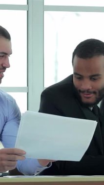 Gösterişli, güneşli bir ofiste, kendine güvenen iki işadamı dinamik bir tartışmaya girişerek, odaklanmış yoğunlukta önemli belgeleri gözden geçiriyorlar. Resmi kıyafetleri ve nişanlı ifadeleri ekip çalışmasından ve stratejik planlamadan oluşan canlı bir atmosfer yaratıyor.