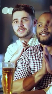 Birinin kısa sakallı, diğerinin kısa kesilmiş saç stili olan iki genç iş adamı, favori futbol takımları için tezahürat yaparken heyecanlı bir anı paylaşıyor ve altın bira bardaklarını şen şakrak kaldırıyorlar. Bar onunla uğulduyor.