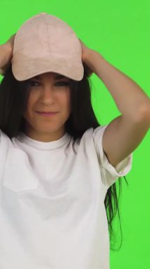 Şık, genç bir esmer kız pembe beyzbol şapkasını neşeli bir şekilde ayarlıyor, canlı yeşil bir arka plana karşı kendine güven yayıyor. Bu modaya uyan genç, günlük beyaz tişörtler giymiş, gençlik enerjisini ve modern yeteneği somutlaştırıyor, izleyicileri,