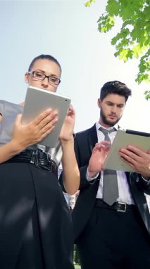 Kendinden emin bir işadamı ve kararlı bir iş kadını, modern mimari ve yemyeşil ağaçlar tarafından çerçevelenmiş canlı bir şehir parkında tabletleri üzerinde yoğun bir işbirliği içinde çalışmaktadırlar. Odaklanmış ifadeleri ve şık profesyonel kıyafetleri yenilik yayıyor.