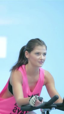 Parlak ışıklarla dolu, hareketsiz bir spor salonunda, atletik kızlar hareketsiz bisikletlerle pedal çeviriyor, enerji ve kararlılık saçıyor. Güçlü bacakları ve odaklanmış ifadeleri bu konuda birbirlerine meydan okurken güçlü bir yoldaşlık bağını yansıtıyor.