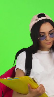 Canlı genç bir kadın neşeli bir telefon sohbeti yaparken neşeli bir şekilde ışıldıyor, renkli ders kitaplarını dengede tutarken modaya uygun bir şapka ve şık güneş gözlüğü takıyor. Canlı bir kromakey arkaplanına göre ayarlayın, bu sahne özetlerName