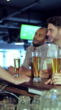 Canlı bir sahnede, her biri büyük ekranda en sevdikleri futbol takımı için tezahürat yaparken kahkaha ve heyecanı paylaşan dört genç adam görülüyor. Parlak bar atmosferi, seslerle dolu.
