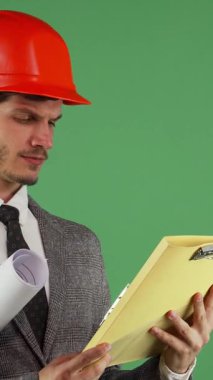 Parlak turuncu bir başlık takan yakışıklı sakallı erkek mühendis, parlak yeşil krom anahtar arka planına karşı bir dizi inşaat belgesini incelerken şaşkın görünüyor. Bu ilgi çekici görüntü, inşaatta karşılaşılan zorlukları özetliyor.