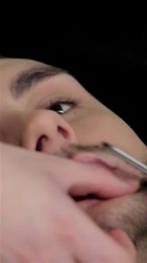 Şık bir genç adam lüks, kaliteli bir berber koltuğunda uzanmış yetenekli bir berberin eski moda tıraşının tadını çıkarıyor. Nostaljik ambiyans zengin ahşap aksanlar ve tıraş köpüğünün aromatik kokusu sayesinde mükemmel bir karışım yaratıyor.