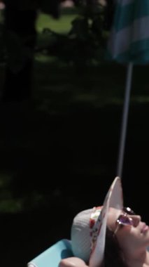 İki muhteşem beyaz kadın, biri esmer, diğeri ateşli kızıl saçlı, ışıl ışıl parlayan bir havuzun kenarında canlı güverte sandalyelerinde uzanıyorlar. Şık bikiniler ve geniş kenarlı şapkalar giyip, rahatlama ve neşe saçarak, sıcacık havayı emerler.