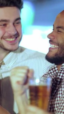Heyecanlı iki arkadaş, beyaz bir adam ve siyah bir adam canlı bir barda neşeli bir şekilde bira bardaklarını kaldırıyor ve ekranda heyecan verici bir futbol maçını kutluyorlar. Bulaşıcı gülüşleri ve coşkulu tezahüratları heyecan verici bir atmosfer yaratıyor.