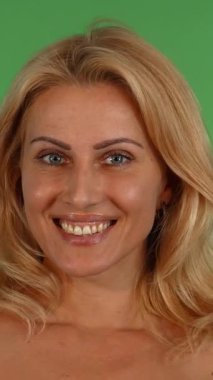 Çekici, olgun, dalgalı sarı saçlı bir kadın ellerini şakaya vurarak yüzünü kapatıyor, canlı yeşil renkli bir arka plana karşı canlı bir duygu yelpazesi sergiliyor. Bu büyüleyici portre, izleyicileri onun mutluluk duygularıyla bağlantı kurmaya davet ediyor.