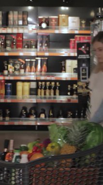 Mutlu bir çift canlı bir hipermarket koridorunda renkli alkollü içecekler seçerken alışveriş arabaları taze ürünlerle dolup taşıyor. Genç beyaz erkek ve kadın neşe saçıyor. Parlak gülümsemeleri heyecanı yansıtıyor.