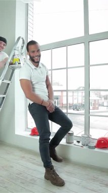 İki hevesli genç müteahhit, biri kendine güvenen bir merdivene tünemiş, diğeri de yanında duruyor, gururla başparmaklarını gösterirken boya kutularını tutuyorlar. Canlı kaskları ve günlük kıyafetleri parlak, havadar, kapalı alana karşı patlar.