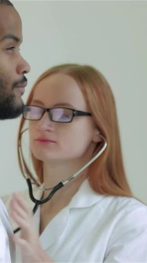 Parlak, modern bir klinikte, kızıl saçlı odaklanmış bir kadın doktor erkek hastalarının kalp atışlarını stetoskop kullanarak dikkatle dinliyor ve besleyici bir sağlık ortamını somutlaştırıyor. Bu samimi an hayati bağlantıyı vurguluyor.