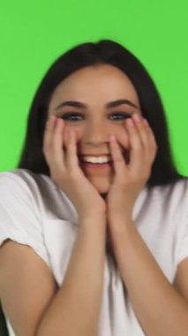 Çekici, genç, esmer bir kadın neşe saçıyor, elleri şen şakrak bir sürpriz jesti ile yüzünü çerçeveliyor. Canlı yeşil bir arkaplan karşısında neşeli yüzü sevinçle dolup taşar ve izleyicileri bulaşıcı bir şekilde paylaşmaya davet eder.