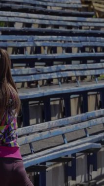 Kararlı genç bir kız, hareketli bir stadyumda heyecan verici bir yarışa hazırlanırken bacaklarına ve ayaklarına odaklanıyor. Renkli koltuklar canlı bir zemin oluşturuyor, atletizm kıyafetlerini vurguluyor ve...