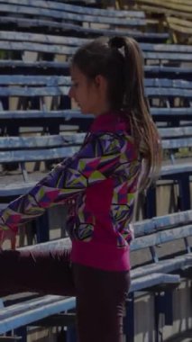 Kararlı genç bir kız dinamik bir bacak esneme hareketine girer, ayağını renkli bir spor kıyafetiyle kaldırır, boş stadyum tribünlerinin arka planına kurulur. Bu canlı sahne enerji ve odaklanma yayıyor. Kendini spora adamış.