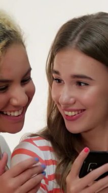 Neşeli kahverengi saçlı beyaz bir kız ve canlı kızıl saçlı bir kız akıllı bir telefonla selfie çekerken keyifli bir anı paylaşıyorlar, yüzleri parlak beyaz bir fon karşısında kahkahalarla parlıyor. Günlük kıyafetleri ve neşeli ifadeleri.