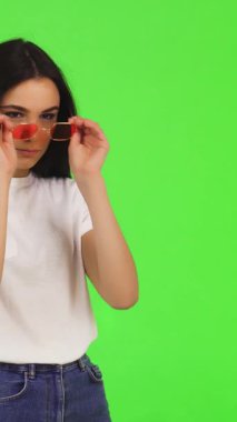 Uzun, dalgalı siyah saçlı çarpıcı genç bir kadın modaya uygun pembe güneş gözlüklerini ayarlıyor ve canlı yeşil bir fon karşısında kendine güven yayıyor. Günlük beyaz tişörtü ve rahat tavırları şık ve davetkar bir atmosfer yaratıyor.