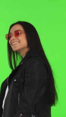 Işıl ışıl genç bir kadın şık alışveriş çantalarını canlı yeşil renkli bir arka planda sergilerken mutluluk saçıyor. Şık bir deri ceket ve modaya uygun pembe güneş gözlükleriyle, gençlik coşkusunun ve modern