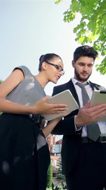 Kendine güvenen bir işadamı ve başarılı bir iş kadını canlı yeşillik ve şık modern mimari ile çevrili bir tabletle neşeyle meşgul oluyorlar. Işıldayan gülümsemeleri ve coşkulu jestleri takım çalışmasının ve başarının heyecanını yayıyor.