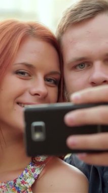 Neşeli genç bir çift, dalgalı kızıl saçlı bir kadın ve modaya uygun sakallı bir adam, güneşli bir bankta otururken akıllı telefonlarıyla bir selfie çekiyorlar. Işıl ışıl gülüşleri yemyeşil bir zemin üzerinde parlıyor.