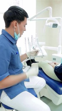 Kendini işine adamış, şık mavi üniformalı genç bir dişçi, gösterişli bir dişçi koltuğunda rahat bir şekilde oturan bir hastayla konuşurken sıcak bir gülümsemeyle öne eğiliyor. Parlak, çağdaş klinik, ışıldayan diş aletleriyle profesyonelliği yayıyor.