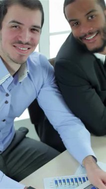 Güneşli modern bir ofiste, biri kıtır mavi gömlekli, diğeri de keskin takım elbiseli iki başarılı iş adamı, başparmak kaldırırken gururla ışıldıyor. Canlı gülüşleri ve kendinden emin tavırları pozitiflik ve takım çalışması yansıtıyor.