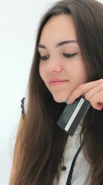 Uzun, dalgalı saçları olan neşeli genç bir kadın kredi kartını tutarken şık, modern dizüstü bilgisayarıyla online alışverişini tamamlamaya hazırlanıyor. Parlak, minimalist arka plan heyecan ve çağdaş yaşam tarzını yansıtıyor.