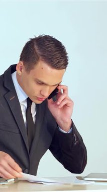 Gösterişli siyah bir takım elbise giymiş kararlı genç bir işadamı, önemli bir telefon görüşmesi yaparken, modern masasındaki önemli belgeleri dikkatle incelerken, kendinden emin bir şekilde gülümsüyor. Parlak ofis ortamı, şık bir dizüstü bilgisayar ve