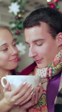 Renkli eşarplara ve rahat kazaklara sarılı neşeli bir çift, sevgi dolu bir şekilde dumanı tüten bir bardağı güzel bir Noel odasında paylaşıyor. Parıldayan ışıkların yumuşak parıltısı ve canlı dekorasyonlar onları sarıp sarmalayarak büyülü bir atmosfer yaratıyor.