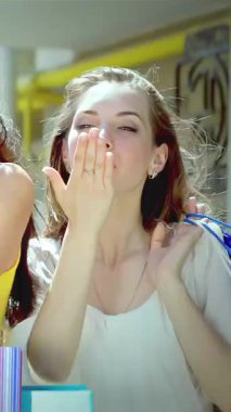 Üç modaya uygun genç kadın, her biri canlı kıyafetler giyip renkli alışveriş torbalarıyla sarılmış, neşeli öpücükler saçıyor, neşe ve dostluk saçıyor. Parlak gülümsemeleri ve canlı ifadeleri büyüleyici bir atmosfer yaratıyor.