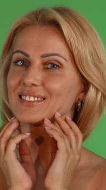 Büyüleyici bir olgun kadın, dalgalı sarı saçları ve çarpıcı mavi gözleri ile kameraya doğru ışıldıyor, yüzüne nazikçe dokunuyor canlı yeşil renkli bir arka plan. Bu büyüleyici portre güzelliği, özgüveni ve sağlıklı bir güzelliği kutluyor.