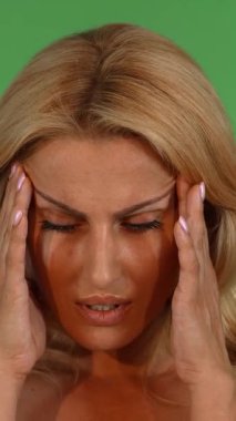 Canlı, yeşil bir krom anahtar stüdyosunda, dalgalı sarı saçlı, çarpıcı olgun bir kadın şakaklarını nazikçe ovuyor, yüz ifadesi, yıpratıcı bir baş ağrısına karşı verdiği mücadeleyi ortaya koyuyor. Zarif özellikleri ile görünür arasındaki zıtlık.