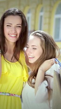 İki şık kız, biri parlak sarı bir elbise içinde, diğeri şık bir bluz içinde, neşeyle sarılıyorlar, renkli alışveriş torbaları. Işıl ışıl gülüşleri ve bulaşıcı kahkahaları canlı bir atmosfer yaratıyor, özlerini mükemmel bir şekilde yakalıyor.