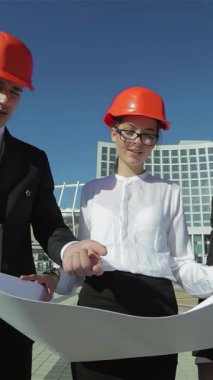 Üç kendine güvenen mimar, şık takımlar ve parlak turuncu kasklar giyip, açık havada detaylı bir iş planını tutkuyla gözden geçiriyorlar. Odaklanmış ifadeleri ve işbirlikçi enerjileri, yenilikçi dönüşümü öngördüklerinde heyecan yayıyor.