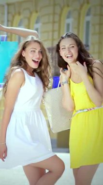 İki ışıl ışıl kız, biri akıcı beyaz elbiseli, diğeri parlak sarı elbiseli, renkli alışveriş torbalarıyla canlı bir alışveriş merkezinin arka planına poz veriyorlar. Bulaşıcı gülümsemeleri ve şakacı jestleri dostluk ruhunu somutlaştırıyor.