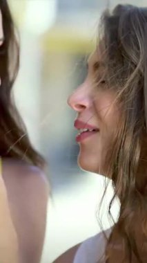 İki neşeli kız, biri uzun siyah saçlı ve diğeri dalgalı kahverengi bukleli, yakından eğiliyor, kalabalık bir alışveriş bölgesinde sırlarını fısıldıyor. Parlak gülümsemeleri ve canlı kıyafetleri arkadaşlığın ruhunu somutlaştıran renklerle doludur.