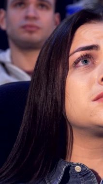 Uzun, dalgalı kahverengi saçlı, çarpıcı beyaz bir kadın loş ışıklı bir tiyatroda büyülenmiş bir şekilde oturuyor. Ekrandaki yoğun dramayı izlerken ifadesel yüzü duyguların kasırgasını gösteriyor. Yakın plan, gözyaşı dolu gözlerini ön plana çıkarıyor.