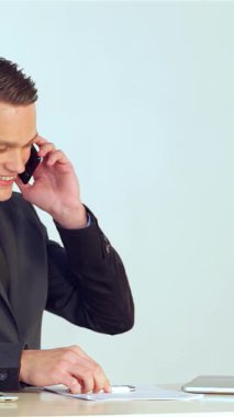 Özel dikim takım elbiseli gülümseyen genç bir işadamı cep bilgisayarına veri girerken aktif olarak bir telefon görüşmesi yapıyor. Enerjik enerjisi ve profesyonel tavırları verimli ve davetkar bir atmosfer yaratıyor.