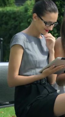 Üç kendine güvenen iş kadını şık bir park bankında oturmuş cihazlarıyla etkileşim halindeyken fikirleri tartışıyorlar. Şık kıyafetleri ve ışıl ışıl gülümsemeleri yemyeşil bir zemin üzerinde canlı ve işbirlikçi bir ruhu temsil ediyor.