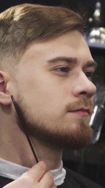 Özenle hazırlanmış sakallı şık bir genç adamın yakın çekimi, yetenekli bir berberin ustalıkla sakalını makasıyla şekillendirmesi gibi ışıldıyor. Bu samimi sahne, özbakım ve erkeksiliği yansıtıyor. Tımarcılığın sanatsallığını vurguluyor.