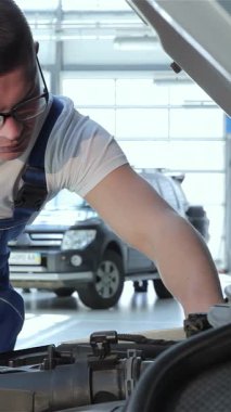 Sarışın, kendini adamış beyaz bir tamirci kaputun üzerine eğilip karmaşık motor parçalarını inceliyor. Mavi tulumlar ve şık gözlükler takmış. Parlak, modern bir garajda profesyonelliği temsil ediyor.