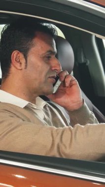 Koyu renk saçlı keyifli bir adam şık, modern bir arabada oturup telefonuyla sohbet ediyor. Bej rengi süveteri zarif kahverengi iç kısmıyla zıtlaşıyor. Gelişmişlik ve neşe saçıyor.