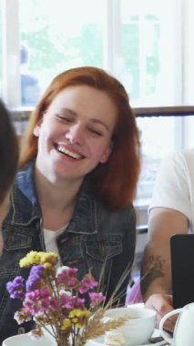 Neşeli, kızıl saçlı genç bir kadının hoş bir kahvaltı eşliğinde yakışıklı erkek arkadaşı ve yakın arkadaşıyla kahkahalarını paylaşması. Sıcak atmosfer, taze hamur işleri ve zengin kahveyle dolu.