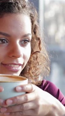 Kıvırcık saçlı neşeli genç bir kadın sıcacık bir kafede sıcak bir fincan kahve içerken mutluluk saçıyor. Yumuşak ışık ve enfes aromalarla dolu davetkar atmosfer, onun mutluluk ve rahatlama anını ve bağlantısını arttırır.