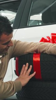 Kendinden geçmiş adam, çarpıcı kırmızı bir fiyonk ile güzelce sarmalanmış iki yeni araba tekerleğinin yanına neşe saçıyor. Elinde onayla ve araba anahtarlarıyla, lüks bir otomobil galerisine karşı, heyecan verici bir satın alma sevincini yayıyor.