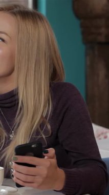 Hoş, genç, beyaz, dalgalı sarı saçlı bir kadın canlı bir kafede oturuyor. Akıllı telefon elinde, hayallerinde kaybolmuş. Sıcak, cezbedici bir dekor ve büyüleyici gülüşü huzurlu bir atmosfer yaratıyor, yansıma anları için mükemmel.