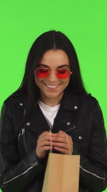 Uzun siyah saçlı ve son moda kırmızı güneş gözlüklü neşeli genç bir kadın, parlak yeşil renkli bir arka plana sahip alışveriş çantasına bakarken heyecan saçıyor. Bu dinamik sahne perakende terapisinin coşkulu heyecanını yakalıyor.